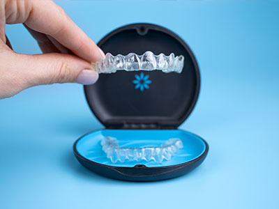 The image shows a person s hand holding a clear plastic tray containing a set of transparent dental aligners, with an open blue case displaying one of these aligners in front.