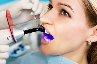 A woman receiving dental treatment with a device that appears to be an oral scanner or 3D printer, while wearing a surgical mask and seated in a dental chair.