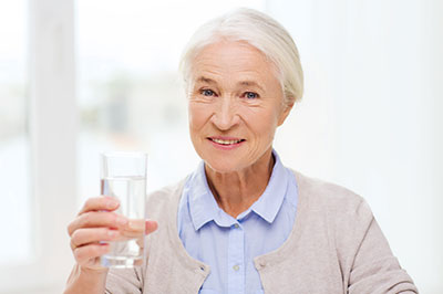 The image shows an elderly woman holding a glass of water with both hands, smiling gently at the camera.