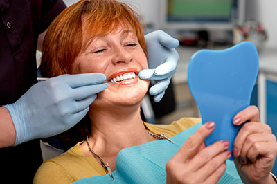 The image shows a woman seated in a dental chair with a smile on her face, holding a blue dental impression tray, while a dental professional is adjusting her mouth with a mirror and a dental tool.