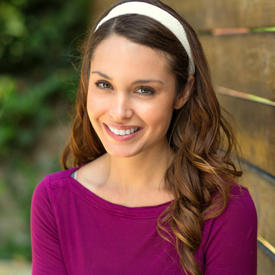 A smiling woman with long hair wearing a purple top and a headband poses for a portrait against a wooden fence.