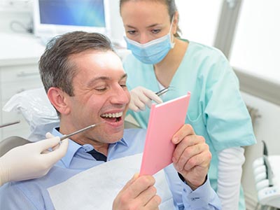 The image shows a man sitting in a dental chair with a smile on his face, holding up a pink card with a surprised expression while looking at it. In the background, there s a female dental professional wearing gloves and a mask, standing behind him, smiling as well.