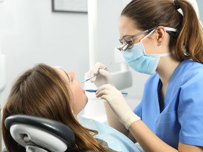 A dental hygienist performing a cleaning procedure on a patient s teeth while wearing protective gloves and a mask, with a woman seated in the dental chair receiving the service.