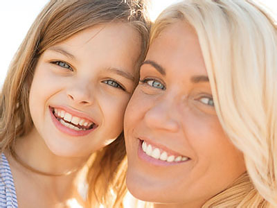 A woman and young girl are smiling at the camera  they appear to be enjoying a sunny day outdoors.