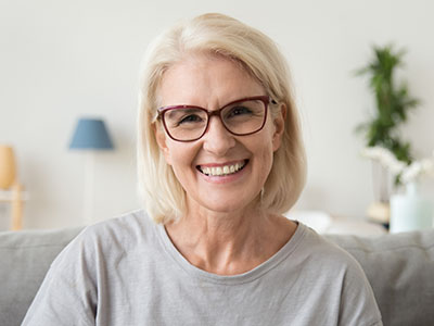 The image features a woman with blonde hair, wearing glasses, smiling at the camera. She has short hair and appears to be indoors with a light-colored wall behind her.