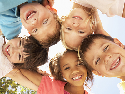 The image shows a group of children of varying ages posing together with smiles, standing outdoors under bright sunlight.