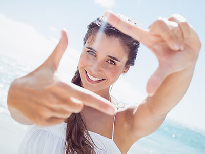 A woman taking a selfie with her hand while smiling at the camera against a beach backdrop.